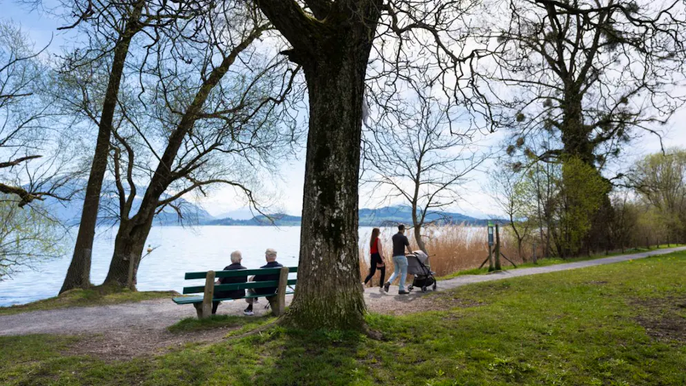 Zuger Lorzenebene ist die Landschaft des Jahres 2026 – Gallery. Alte Menschen sitzen auf einer Bank, während junge Menschen am Seeufer des Zugersees auf der Lorzenebene spazieren.
