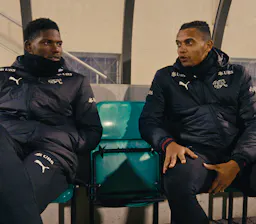 Die Schweizer Nationalspieler Breel Embolo und Manuel Akanji sitzen in Trainingsjacken der Nationalmannschaft auf einer Stadion-Sitzbank und unterhalten sich während einer Pause.
