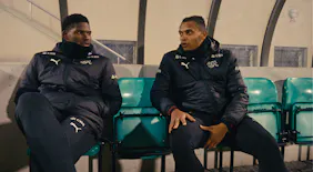 Die Schweizer Nationalspieler Breel Embolo und Manuel Akanji sitzen in Trainingsjacken der Nationalmannschaft auf einer Stadion-Sitzbank und unterhalten sich während einer Pause.