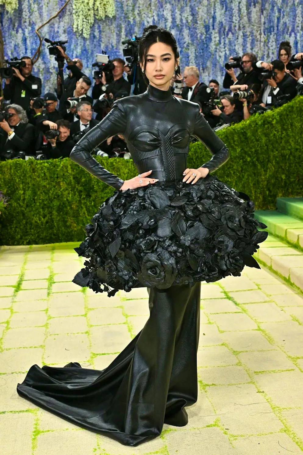 MET Gala - les looks du tapis rouge. Jung Ho-yeon, mannequin sud-coréen, signe une apparition impressionnante dans une robe en cuir intégral, dotée d’un corset intégré et d’un peplum exagéré orné de fleurs en trois dimensions, pour une accentuation maximale des hanches. Un look qui mise clairement sur la structure, la rigueur et une silhouette hautement dramatique.