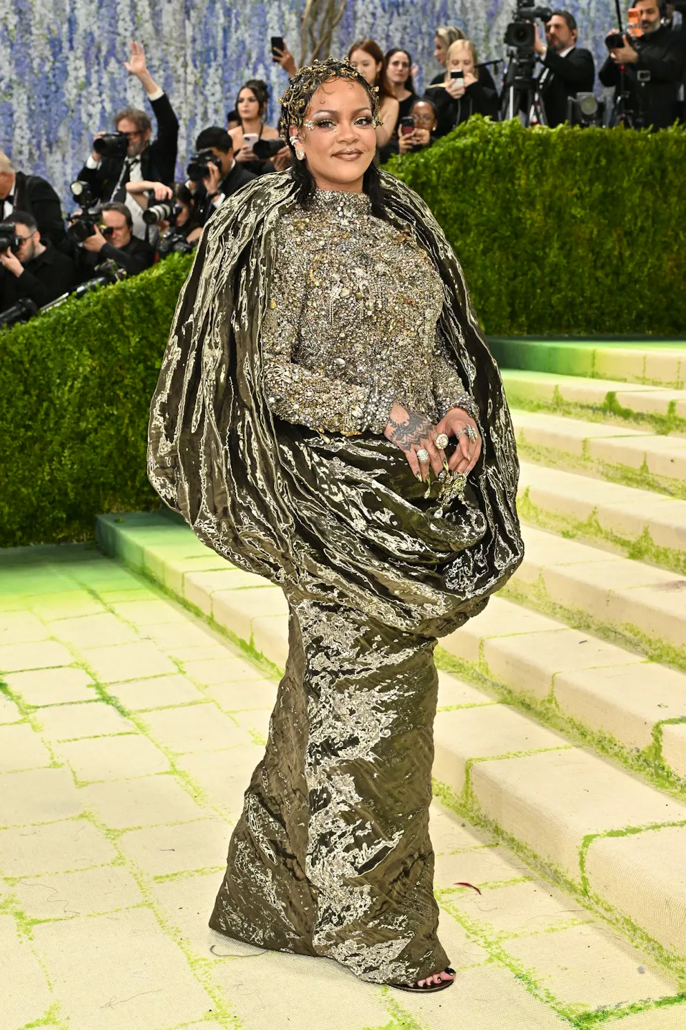 Rihanna setzt auf einen Metallic-Look – und erneut auf maximales Volumen dank opulenter Drapierungen. Doch angesichts der hunderttausenden Kristallperlen, antiken Juwelen und Ketten, die ihr Oberteil überziehen, wirkt «metallic» fast ein wenig… untertrieben.