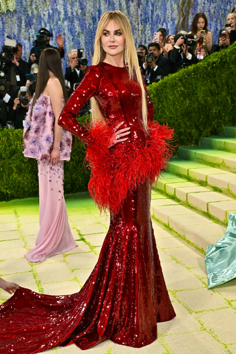 MET Gala - les looks du tapis rouge. Un clin d’œil à la malle de carnaval ? Nicole Kidman n’hésite en tout cas pas à mêler des éléments voyants comme les paillettes et les plumes, le tout dans un jeu de rouges ton sur ton. Pourtant, elle parvient à rendre l’ensemble élégant, notamment grâce à une silhouette fermée qui dévoile très peu de peau. Ses cheveux, quant à eux, sont lissés avec une sobriété minimaliste.