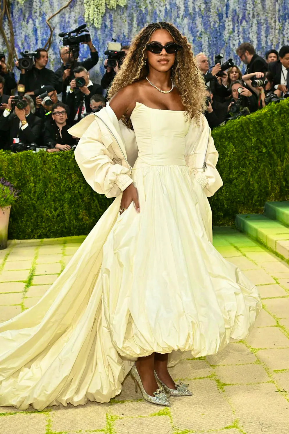 MET Gala - les looks du tapis rouge. Blue Ivy Carter fait exception à l’excentricité ambiante et opte pour un look nettement plus portable : elle porte la jupe ballon tendance en version midi, assortie d’un manteau coordonné, le tout dans un doux jaune pastel. Elle complète la tenue avec des chaussures scintillantes, des ondulations naturelles et des lunettes de soleil. Un look qui évoque davantage un quotidien stylé qu’un costume de scène.