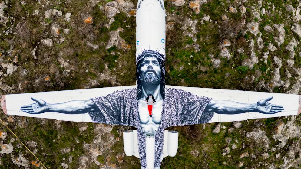 Der Berner Künstler Vierwind alias Micha Haeni in roter Hose auf seinem Kunstwerk: der gekreuzigte Christus, gemalt auf auf ein Flugzeug und betrachtet aus der Himmelsperspektive. Für Haeni symbolisiert es, dass an Himmelfahrt Gott herabsteigt zu den Menschen.