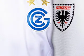 Die Vereinslogos von GC Zürich und dem FC Aarau stehen im Fokus. Das Bild zeigt die Trikots beider Teams vor dem Duell in der Super League. Nahaufnahme der Trikots von GC Zürich und dem FC Aarau mit den beiden Vereinslogos nebeneinander.