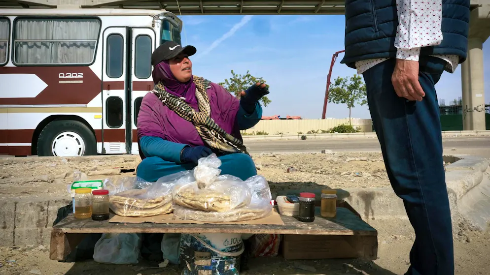 Lunch break at the construction site: In the dusty desert wind, a woman on the roadside sells fresh flatbread and halawa to workers. While cranes stand still in the background, construction workers grab a quick meal here - in the middle of the sand, between heat and concrete.