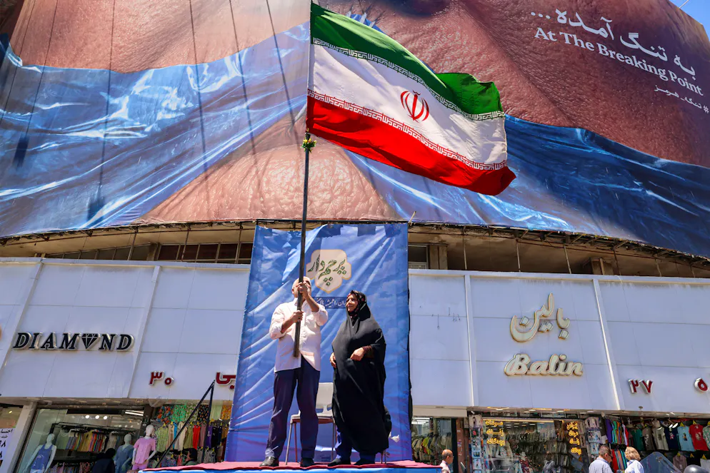 Un homme brandit un drapeau iranien devant un panneau politique représentant le président américain Donald Trump et le détroit d’Ormuz, sur la place Valiasr à Téhéran, le 26 mai 2026. Dans la nuit, les forces américaines ont mené des frappes contre des sites de missiles iraniens et des embarcations accusées de vouloir poser des mines dans le Golfe.