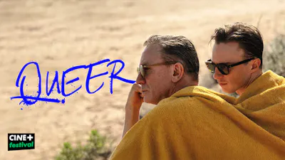 Une affiche au format paysage montrant deux hommes (Daniel Craig et Drew Starkey) assis côte à côte en extérieur, enveloppés ensemble dans une grande serviette ou couverture jaune moutarde. Ils portent tous les deux des lunettes de soleil et regardent vers la droite. Sur la gauche, le mot 