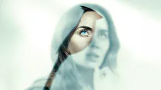 Nahaufnahme einer Frau mit stechend blauen Augen, die durch einen Riss in einer fahlblauen, Nebel-ähnlichen Schicht nach oben blickt.