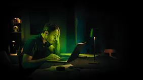 Ein erschöpfter Mann sitzt nachts in einem dunklen Raum vor seinem beleuchteten Laptop und stützt verzweifelt den Kopf mit der Hand ab
